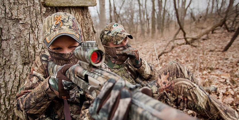A hunter in camoflauge, sitting next to another hunger, takes aim with a firearm.